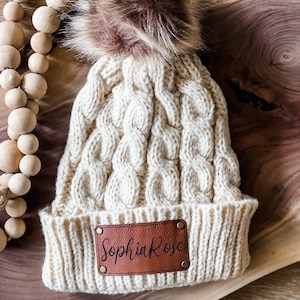 Peut inclure: Un bonnet en tricot crème avec un pompon en fausse fourrure marron. Le bonnet a un patch en cuir marron avec le nom "Sophia Rose" écrit en noir.