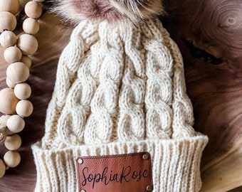 May include: A cream-colored knitted beanie with a brown faux fur pom pom. The beanie has a brown leather patch with the name "Sophia Rose" written in black.