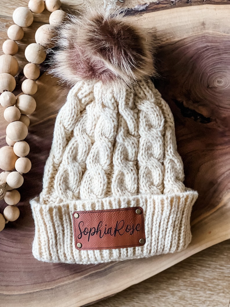 May include: A cream-colored knitted beanie with a brown faux fur pom pom. The beanie has a brown leather patch with the name "Sophia Rose" written in black.