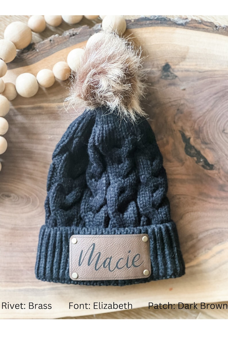May include: Black knit beanie with a faux fur pom pom and a brown leather patch that says "Macie" in a cursive font. The patch has brass rivets.