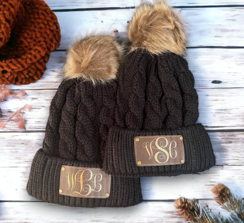 May include: Two black knit winter hats with faux fur pom poms. Each hat has a brown leather patch with gold monogram initials. The initials on the hats are "WLL" and "VSC".