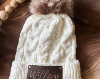 May include: White cable knit beanie with a brown faux fur pom pom and a brown leather patch that says "Willow" in gold lettering.