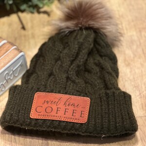 May include: A dark green knitted beanie with a brown faux fur pom pom. The beanie has a brown leather patch with the text "sweet home COFFEE Robertsdale Alabama" on it.