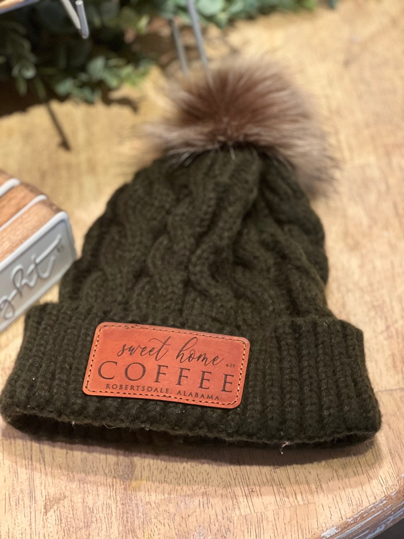 May include: A dark green knitted beanie with a brown faux fur pom pom. The beanie has a brown leather patch with the text "sweet home COFFEE Robertsdale Alabama" on it.