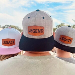 May include: Three gray baseball caps with black brims. The front of each cap has a brown leather patch with the words "LEGEND", "LEGACY", and "LEGACY" on them.