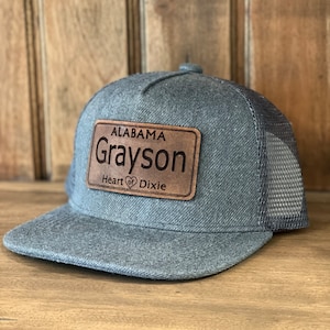 Peut inclure: Un chapeau de camionneur gris et noir avec un patch en cuir marron qui dit "Alabama Grayson Heart of Dixie".
