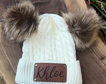 Peut inclure: Bonnet en tricot blanc avec deux pompons en fausse fourrure marron et un patch en cuir marron avec le nom "Khloe" écrit en cursive.