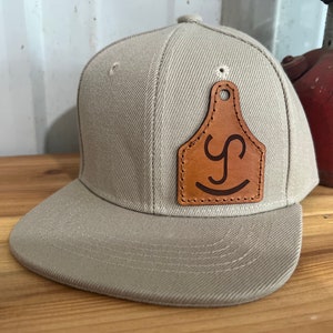 Puede incluir: Una gorra de béisbol beige con un parche de cuero marrón que presenta una letra estilizada "Y" y una cara sonriente. El parche tiene la forma de una etiqueta para el oído del ganado.