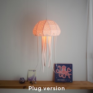 Könnte beinhalten: Eine hängende Lampe in Form einer Qualle mit einem pfirsichfarbenen, geometrischen Schirm, der mit kleinen Löchern versehen ist. Weiße Bänder hängen von unten herab, und der Text "Plug version" ist unten sichtbar. Die Lampe hängt über einem Holzregal.