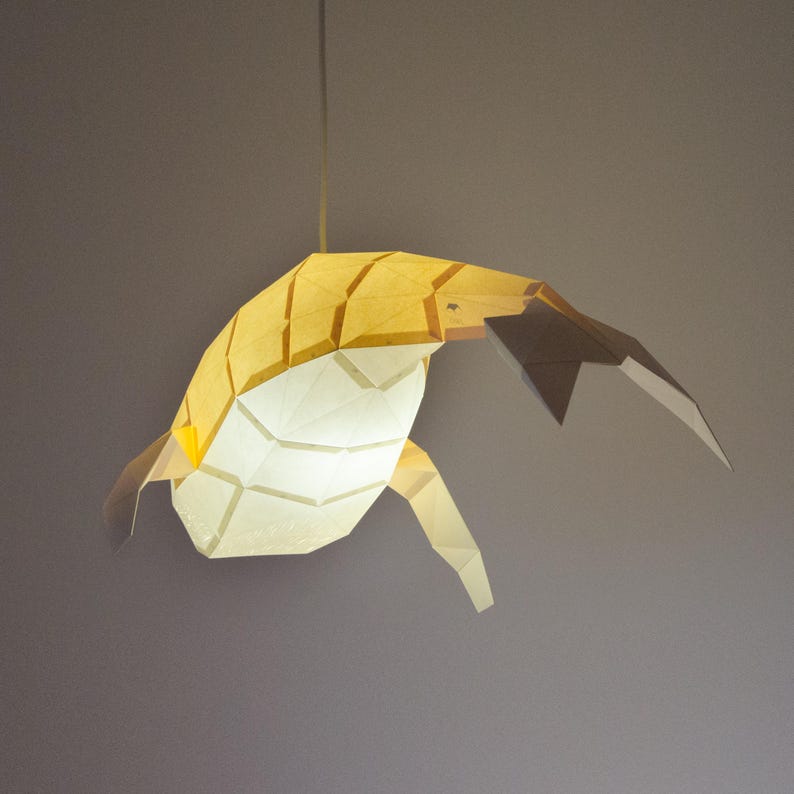 Puede incluir: Una pantalla de l&aacute;mpara de papel en forma de ballena, de color amarillo y blanco, colgada del techo. La ballena est&aacute; suspendida en el aire, con la cola y las aletas extendidas.