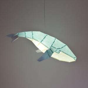 Puede incluir: Una pantalla de l&aacute;mpara de papel en forma de ballena azul claro que cuelga del techo. La pantalla est&aacute; hecha de papel y tiene un dise&ntilde;o geom&eacute;trico.