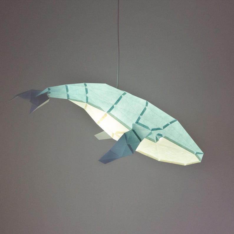 Puede incluir: Una pantalla de l&aacute;mpara de papel en forma de ballena azul claro que cuelga del techo. La pantalla est&aacute; hecha de papel y tiene un dise&ntilde;o geom&eacute;trico.