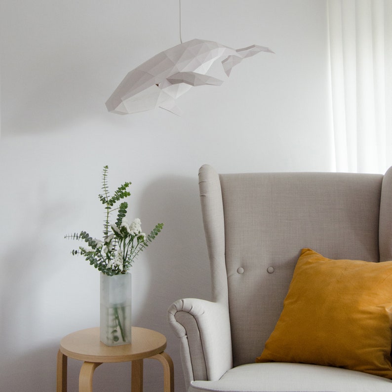 Puede incluir: Un m&oacute;vil de papel blanco en forma de ballena colgando del techo sobre un sill&oacute;n beige con un coj&iacute;n amarillo mostaza. Un jarr&oacute;n con flores blancas se encuentra sobre una mesa auxiliar de madera.