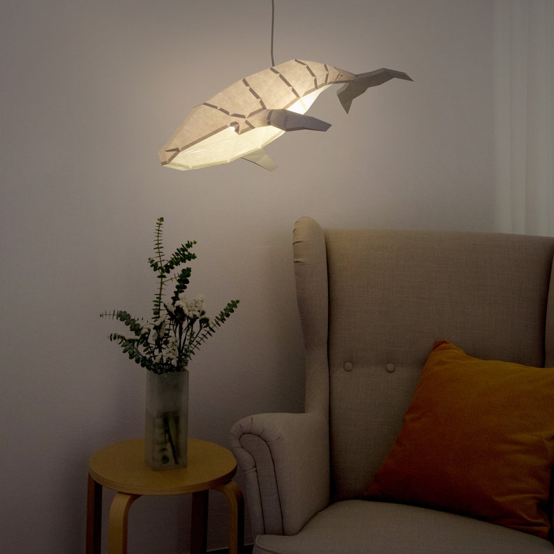 Puede incluir: Una pantalla de l&aacute;mpara de papel blanca con forma de ballena cuelga del techo, iluminando una habitaci&oacute;n con un sill&oacute;n beige y un jarr&oacute;n de flores blancas sobre una mesa de madera.