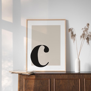 May include: A framed print with a beige background and a black letter "C" in a modern font. The print is on a wooden shelf with a vase of dried flowers.