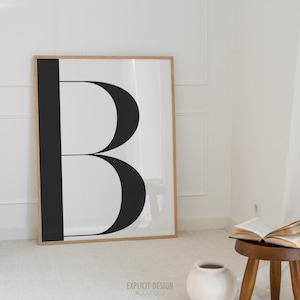 May include: A framed art print featuring a large black letter "B" against a white background. The artwork is in a wooden frame and leans against a white wall. The room has a minimalist aesthetic with a small wooden stool and a vase.