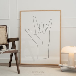 Puede incluir: Un dibujo en blanco y negro de una mano haciendo el signo de lenguaje de señas 'Te amo'. El dibujo está enmarcado en un marco de madera claro y está colgado en una pared blanca.