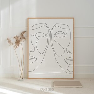 May include: A framed art print with a minimalist line drawing of two faces in black on a cream background, set within a light wood frame. The print is propped against a white wall, with a vase of dried plants and a small book beside it.