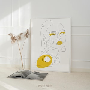 以下が含まれることがあります： 白い背景に、女性の顔のミニマリストな線画が描かれた額入りのアートプリント。 黄色のアイシャドウと口紅が特徴です。 下部には「EXPLICIT DESIGN」の文字があります。