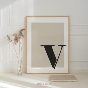 May include: A framed print with a beige background and a large black letter "V" in the center. The frame is a light wood color. The print is sitting on a white surface with a book in front of it.