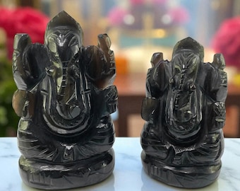 Op de afbeelding: Twee donkergrijze stenen sculpturen van de hindoeïstische godheid Ganesha. De sculpturen zijn gedetailleerd met gebeeldhouwde kenmerken, waaronder de olifantenkop, grote oren en meerdere armen. De beelden worden tentoongesteld op een wit marmeren oppervlak.