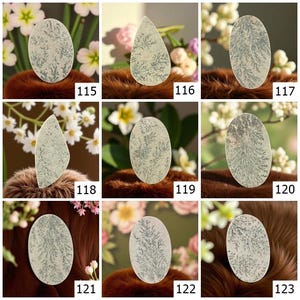May include: A collection of nine dendritic agate gemstones in various shapes. Each stone features a cream-colored base with intricate, fern-like patterns in shades of gray and green. The gemstones are displayed against a backdrop of flowers and brown fur.