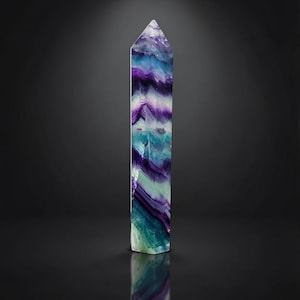 Könnte beinhalten: Ein hoher, polierter Fluorit-Kristallturm mit ausgeprägten, geschichteten Bändern in tiefem Violett, leuchtendem Türkis und durchscheinendem Hellgrün. Der Stein ist in Form eines hexagonalen Prismas mit einer spitz zulaufenden Spitze geschliffen.