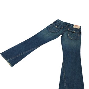 K&ouml;nnte beinhalten: Eine blaue Jeans mit Bootcut-Beinen. Die Jeans hat einen Used-Look und ein kleines rot-wei&szlig;es Label auf der Ges&auml;&szlig;tasche mit dem Schriftzug "True Religion" in Wei&szlig;.
