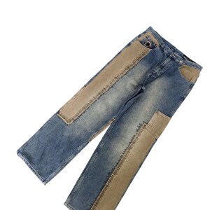 Peut inclure: Un jean en denim bleu avec des empiècements et des coutures en denim marron. Le jean a une coupe décontractée et une jambe droite.