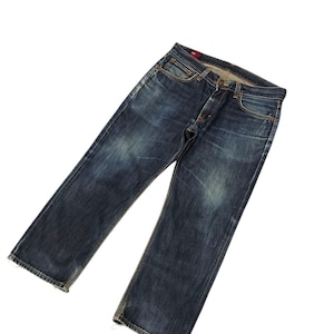 Puede incluir: Un par de vaqueros de mezclilla azul oscuro con un diseño clásico de cinco bolsillos. Los vaqueros tienen bragueta con botones y un aspecto ligeramente descolorido, con sutiles marcas de desgaste. La mezclilla es de color azul oscuro, con zonas más claras en los muslos y las rodillas.