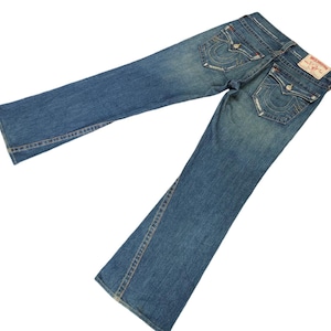 May include: A pair of blue denim jeans with a bootcut leg. The jeans have a light wash and are distressed. The back pockets have a stitched design with the words "True Religion" in white.