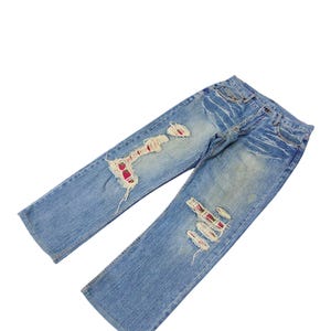 May include: Light blue denim jeans with distressed details. The jeans feature ripped sections with red fabric peeking through. The jeans are laid flat against a white background, showcasing their worn and vintage aesthetic.