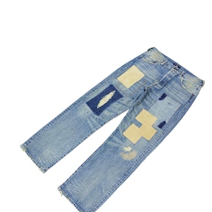 May include: Light blue denim jeans with multiple patches in various shapes and colors. Patches include light beige, dark blue, and white. The jeans have a distressed look with frayed edges and visible wear.