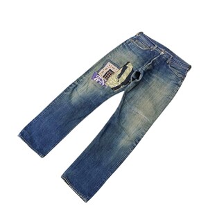 May include: Faded blue denim jeans with a decorative patch on the back pocket. The patch features a combination of patterns and colours, including white, blue, and purple. The jeans have a classic five-pocket design.