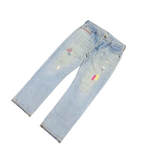 May include: Light blue denim jeans with patches and distressed details. The jeans have a classic five-pocket design, a button and zipper closure, and a straight leg fit. Patches of pink, yellow, and red are visible on the jeans.