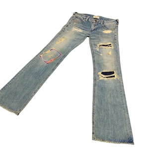 May include: Light blue denim jeans with distressed details, including rips and patches. The jeans have a classic five-pocket design and a relaxed fit. The denim has a faded wash, giving it a worn-in look. The jeans are laid flat against a white background.