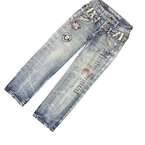 May include: Light blue denim jeans with distressed detailing and patches. The jeans feature a skull patch and text that reads "Signal". The jeans have a classic five-pocket design.
