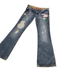 May include: Blue denim flared jeans with decorative patches and distressed details. The jeans feature a light wash with white paint splatters and a faded appearance. Patches include cartoon characters and floral designs.