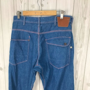 May include: Blue denim jeans with orange stitching. The jeans have large back pockets, a brown leather patch, and a button detail on the back pocket. The jeans are hanging on a wooden hanger.