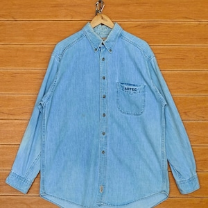 Puede incluir: Camisa vaquera azul claro con botones y mangas largas. La camisa tiene un bolsillo en el pecho con el texto "AZTEC". Cuelga de una percha de madera. El fondo es una pared de paneles de madera.