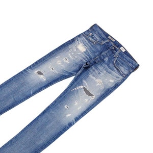 Puede incluir: Un par de pantalones vaqueros de mezclilla azul claro con detalles desgastados y decoloración. Los pantalones vaqueros tienen varias rasgaduras y roturas en la tela.