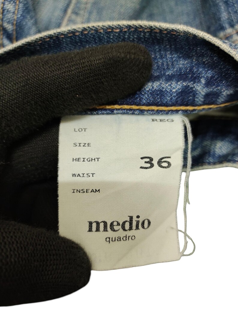 Puede incluir: Primer plano de una prenda vaquera azul claro con una etiqueta blanca. La etiqueta muestra la talla 36 y la marca "medio quadro". El tejido vaquero muestra signos de desgaste.