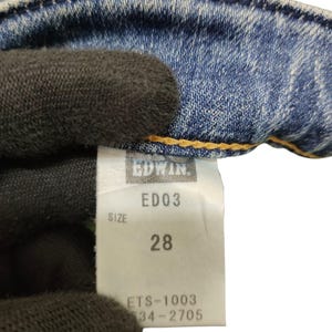 May include: Close-up of a light blue denim jeans tag. The tag reads "EDWIN" and "ED03" with a size of "28". The tag also includes the numbers "ETS-1003" and "34-2705". The jeans are visible in the background.