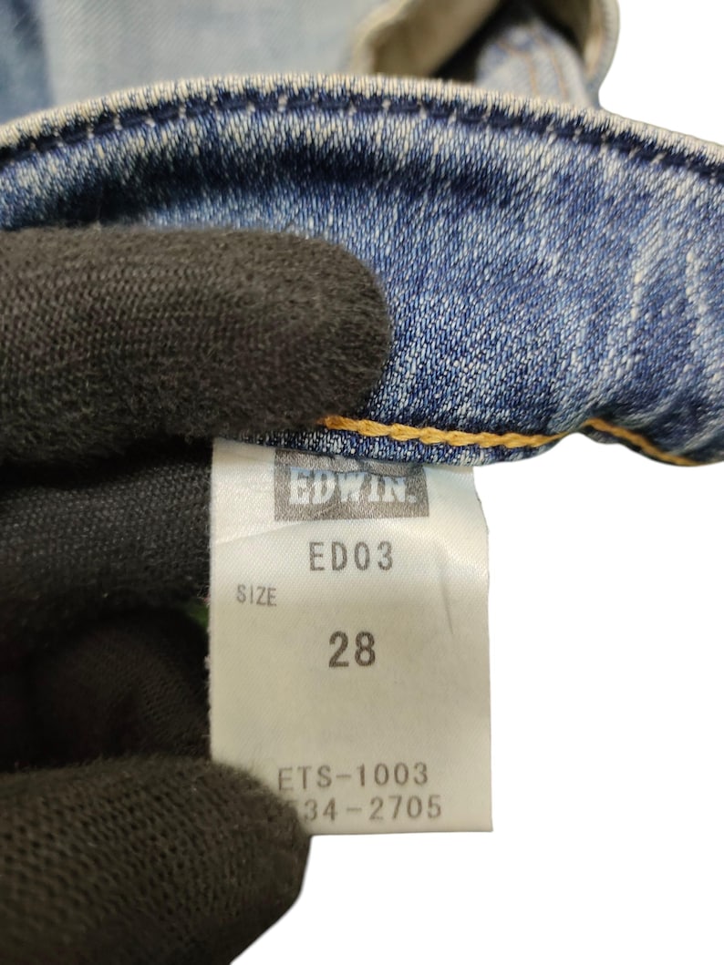 May include: Close-up of a light blue denim jeans tag. The tag reads "EDWIN" and "ED03" with a size of "28". The tag also includes the numbers "ETS-1003" and "34-2705". The jeans are visible in the background.