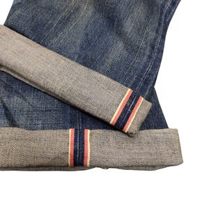 Puede incluir: Primer plano de unos vaqueros de mezclilla azules con un dobladillo vuelto. El dobladillo revela un detalle de rayas rojas, blancas y azules. Los vaqueros tienen un aspecto cl&aacute;sico y desgastado, con textura visible y decoloraci&oacute;n sutil.