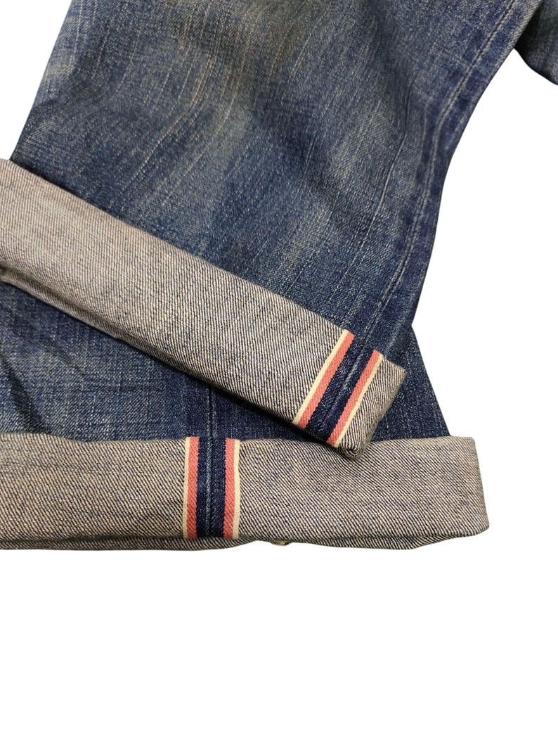 Puede incluir: Primer plano de unos vaqueros de mezclilla azules con un dobladillo vuelto. El dobladillo revela un detalle de rayas rojas, blancas y azules. Los vaqueros tienen un aspecto cl&aacute;sico y desgastado, con textura visible y decoloraci&oacute;n sutil.