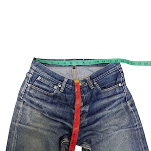 Puede incluir: Vaqueros de mezclilla azul con un lavado descolorido, medidos con una cinta m&eacute;trica verde y roja. La cintura mide aproximadamente 43 cm de ancho. La entrepierna mide aproximadamente 28 cm.