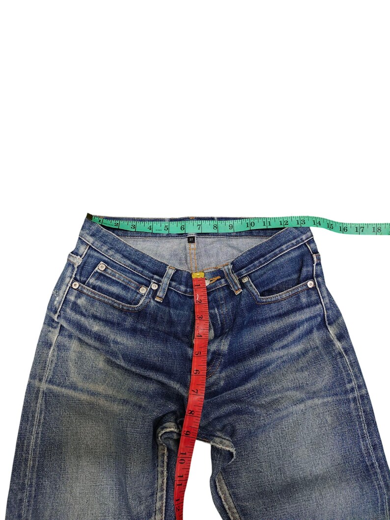 Puede incluir: Vaqueros de mezclilla azul con un lavado descolorido, medidos con una cinta m&eacute;trica verde y roja. La cintura mide aproximadamente 43 cm de ancho. La entrepierna mide aproximadamente 28 cm.