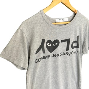 Może przedstawiać: Szara koszulka z kr&oacute;tkim rękawem z czarnym nadrukiem serca z oczami i tekstem "Play Comme des Gar&ccedil;ons"