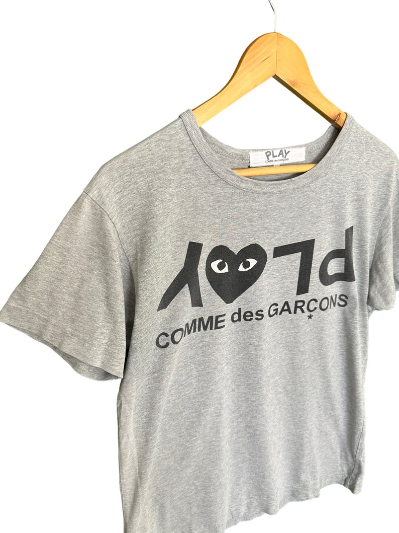 Może przedstawiać: Szara koszulka z kr&oacute;tkim rękawem z czarnym nadrukiem serca z oczami i tekstem "Play Comme des Gar&ccedil;ons"